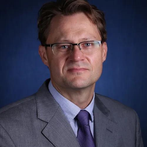 Headshot of person with short hair and glasses wearing a gray suit in front of a dark blue background.