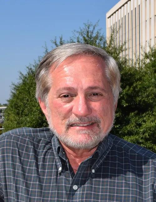 Headshot of person with short hair with beard, wearing a printed button-down shirt with the background of green trees, blue sky and a white building.