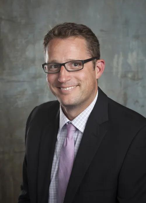 Portrait shot of person with short hair, dark framed eyeglasses wearing a black suit in front of gray background.