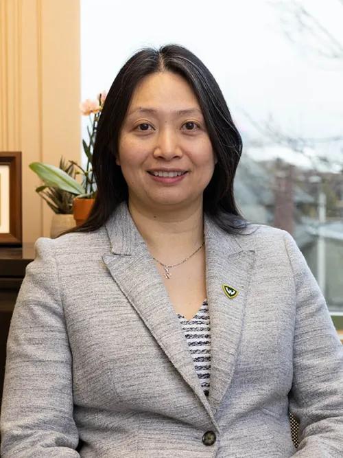 Person with dark, shoulder length hair smiling in gray suit with what appears to be an office setting in the background.