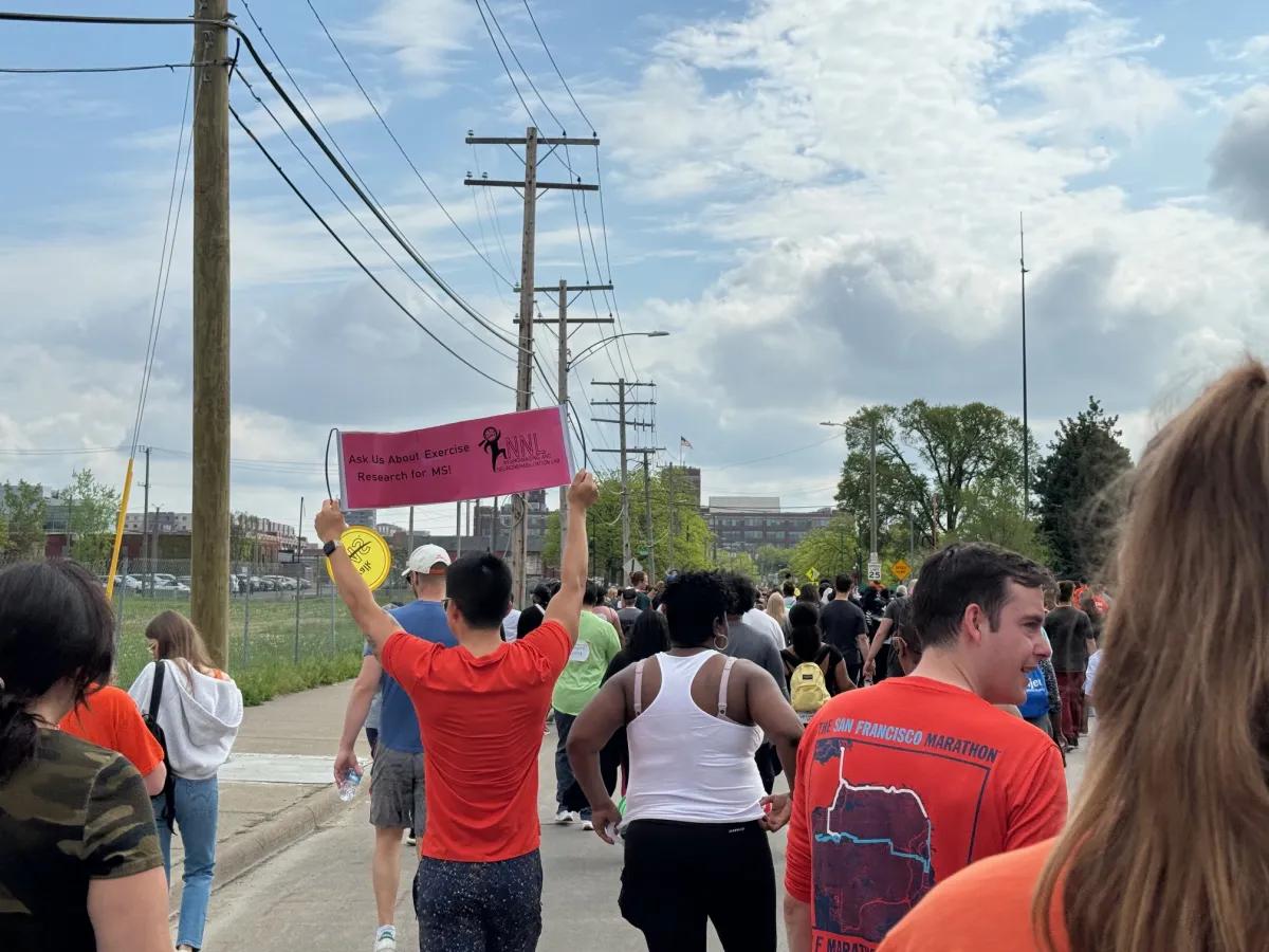 people walking during Walk MS, with one person holding up a sign above their head that says, "Ask Us About Exercise Research for MS!"