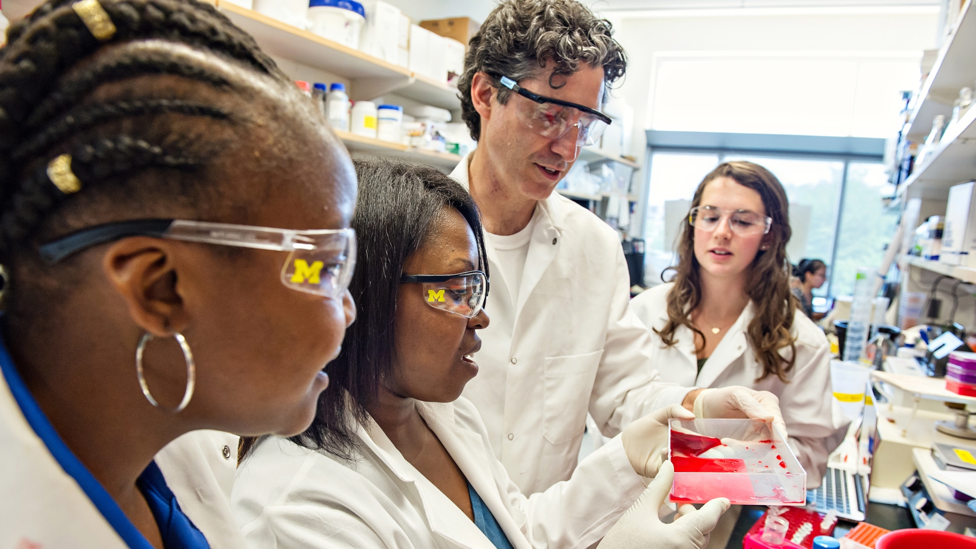 Group of four people in a research lab wearing goggles and white lab coats: one white male, one white female and two black females
