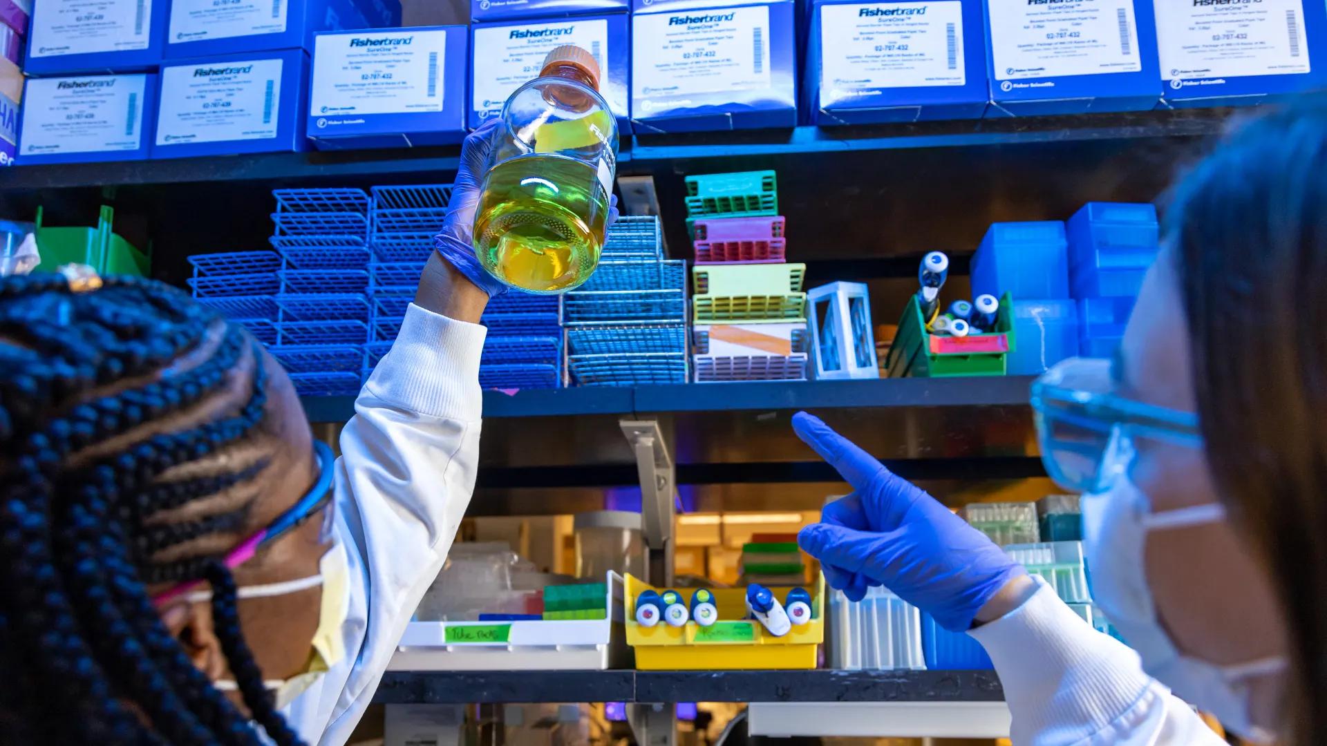 women in lab examining sample jar, one woman points at the sample 