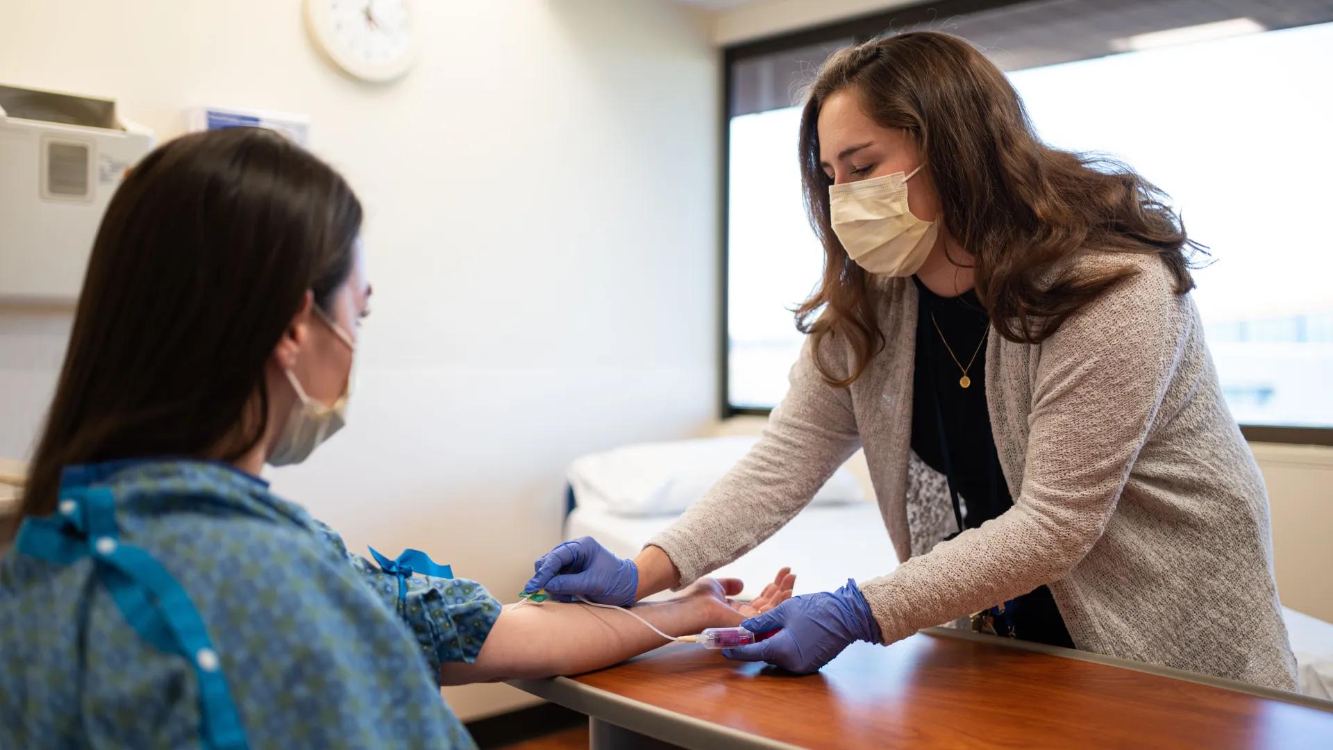 A healthcare worker wearing gloves and a mask is drawing blood from a seated, masked patient in a medical office.
