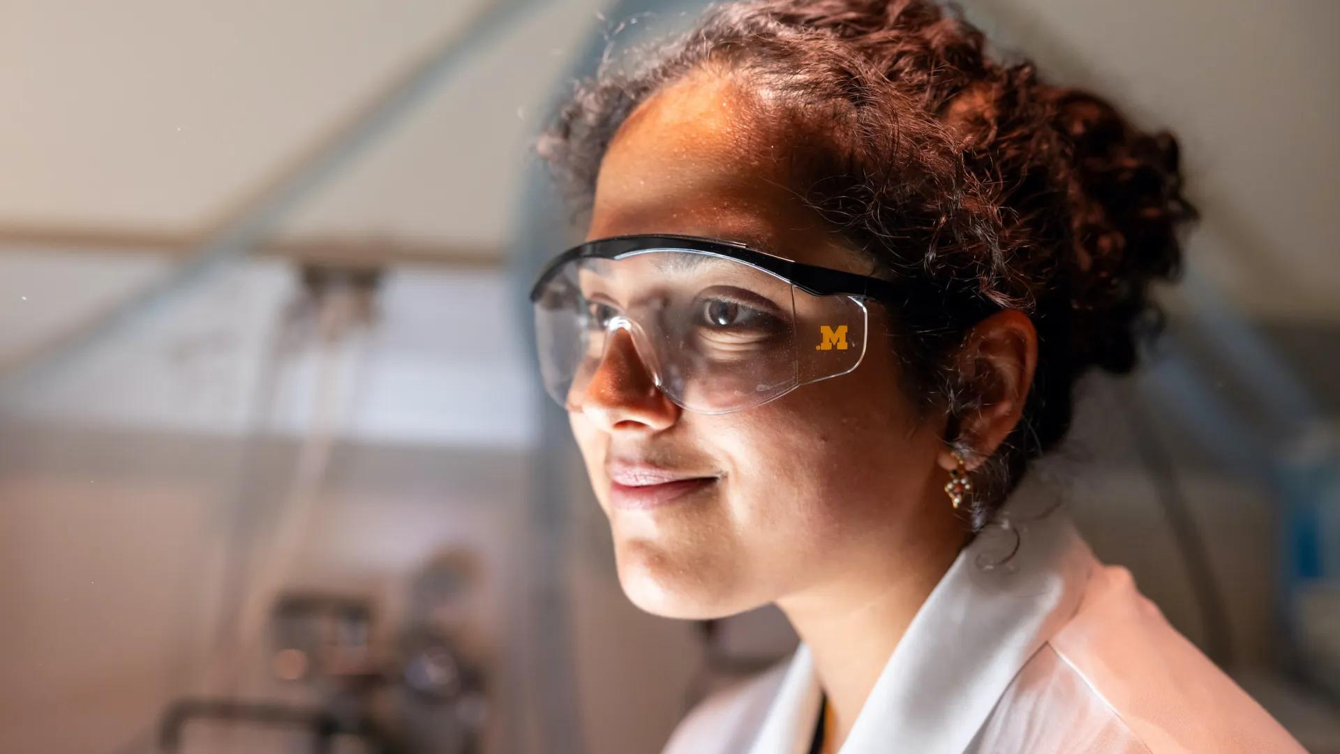 A woman in a lab coat and safety goggles smiles confidently in a laboratory.