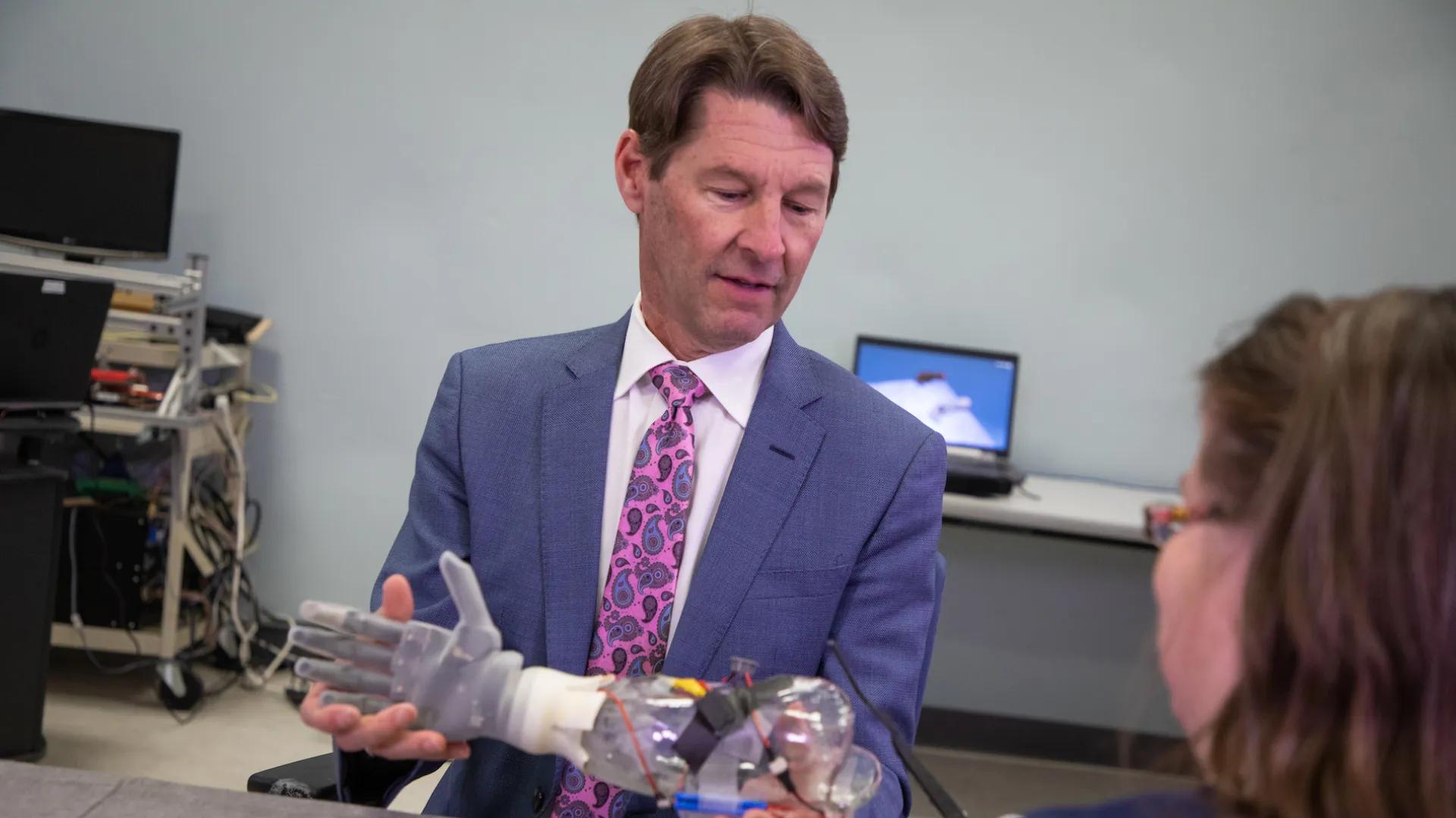 A man in a suit holds a robotic prosthetic hand, explaining it to a seated woman.