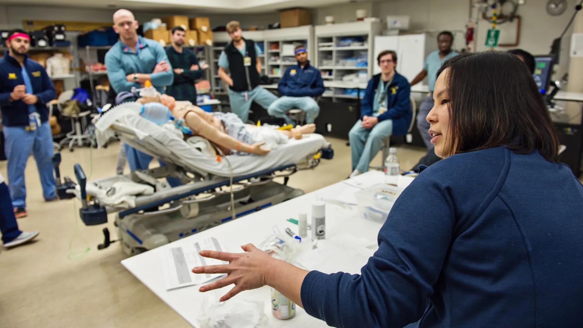 A medical simulation lab scene where a diverse group of people in scrubs attentively observe a woman explaining around a table, next to a patient mannequin on a bed.