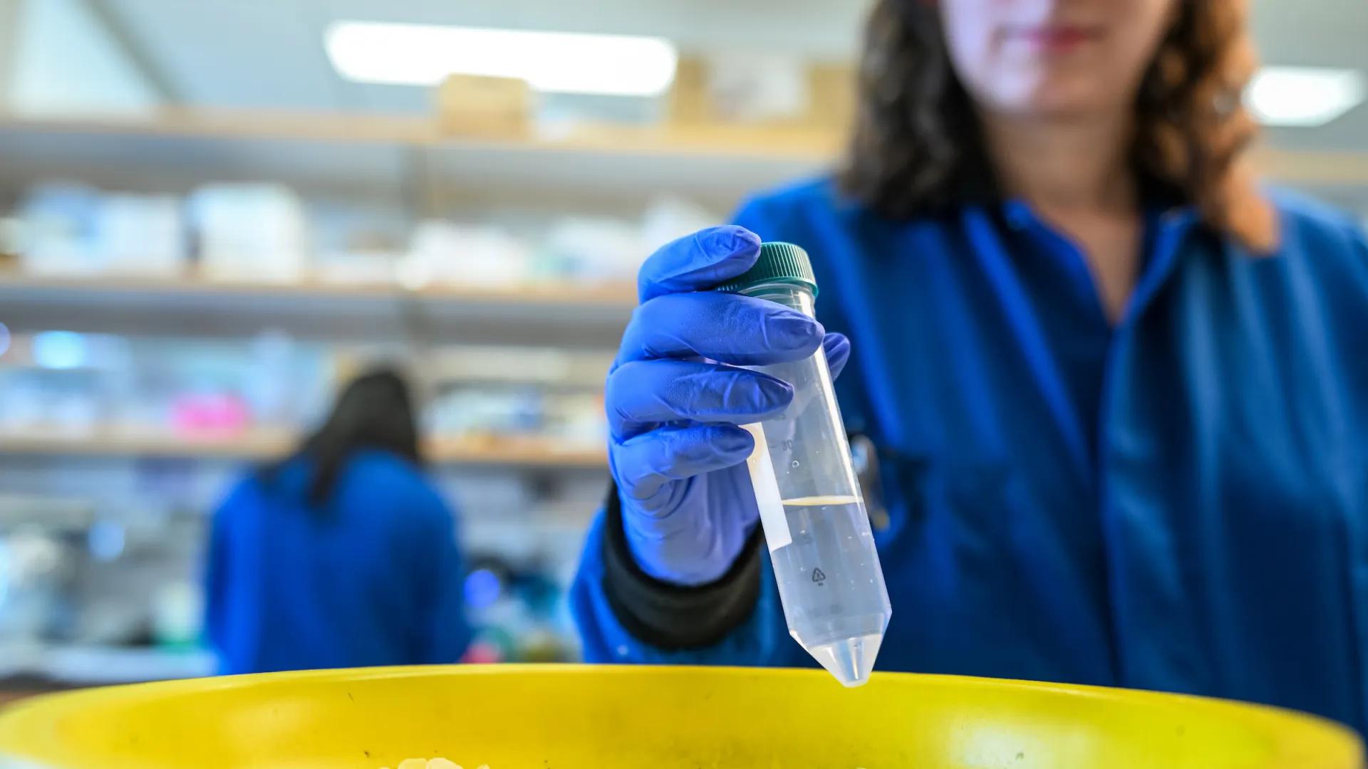 A scientist in a blue lab coat holds a test tube over a yellow bowl filled with ice.