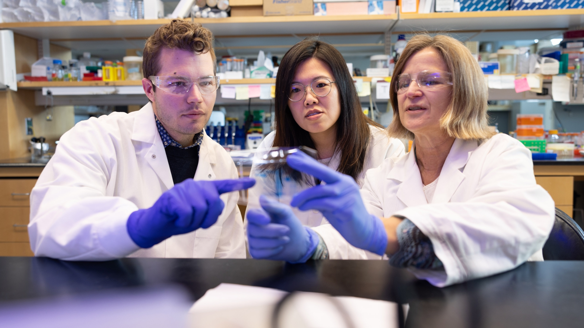 Faculty and students in lab coats examining a specimen under a microscope in a research laboratory.