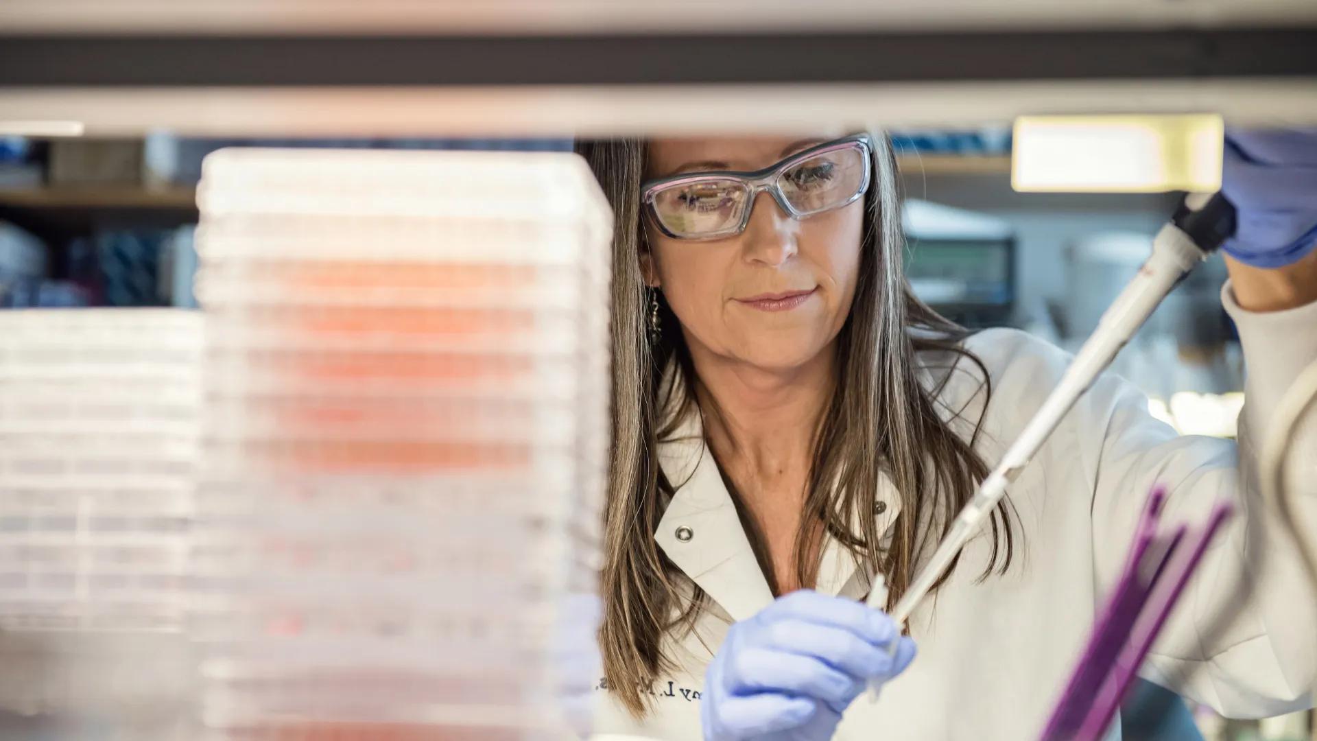 A researcher in a lab coat and safety goggles carefully using a pipette with gloved hands, surrounded by laboratory equipment.