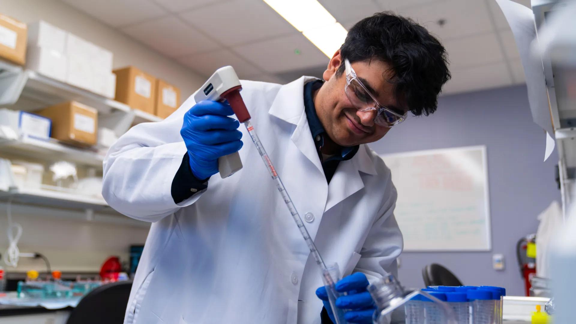A researcher wearing a lab coat, safety glasses, and blue gloves smiles while using a pipette in a laboratory setting.