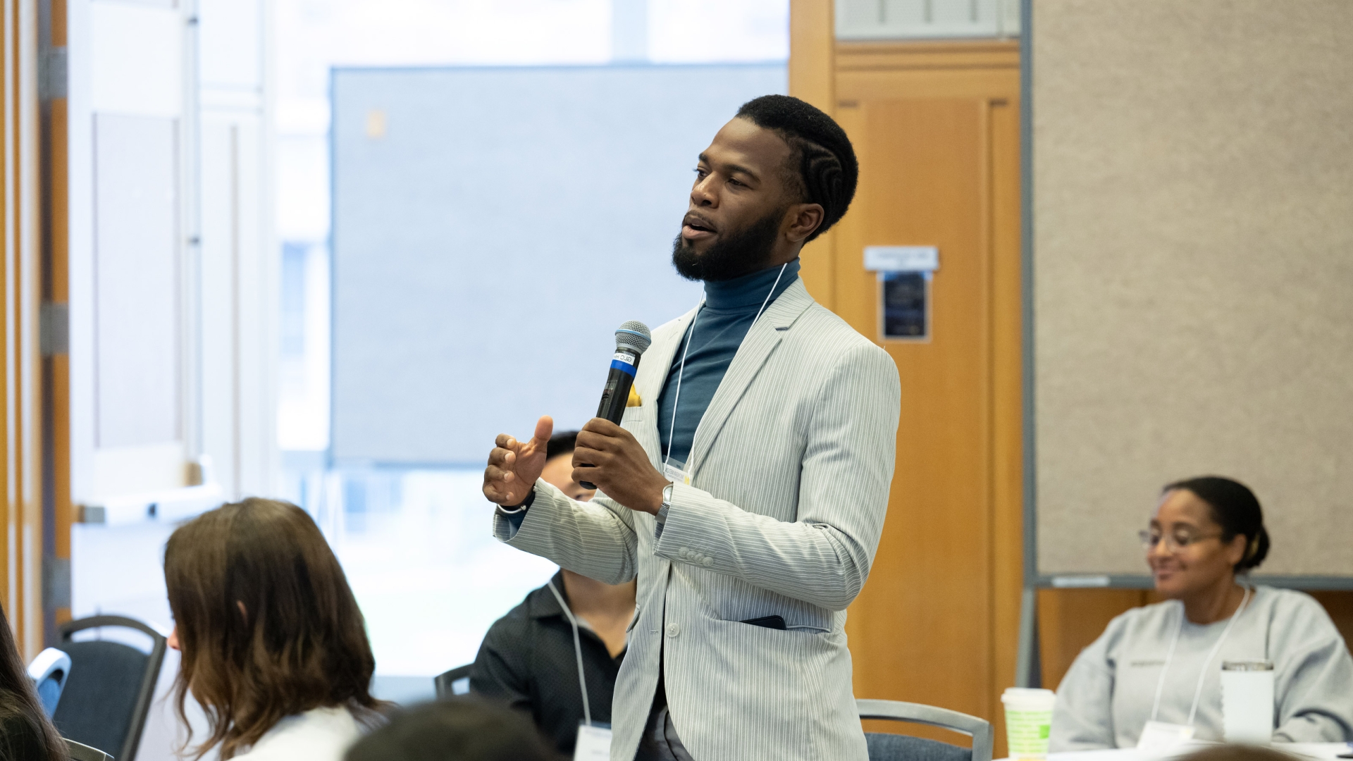 A faculty addresses an audience during a symposium, with attendees seated and listening in the background.