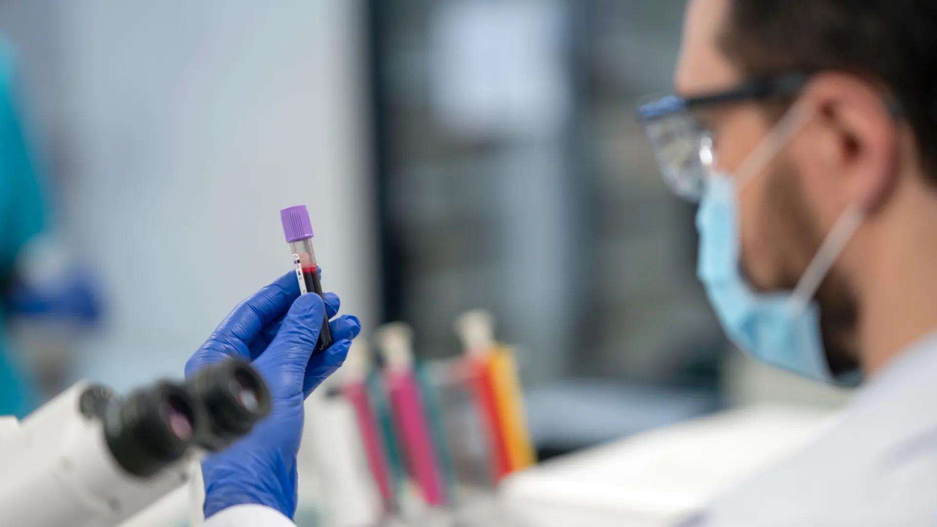 A scientist in a lab coat and mask examines a blood sample in a test tube.