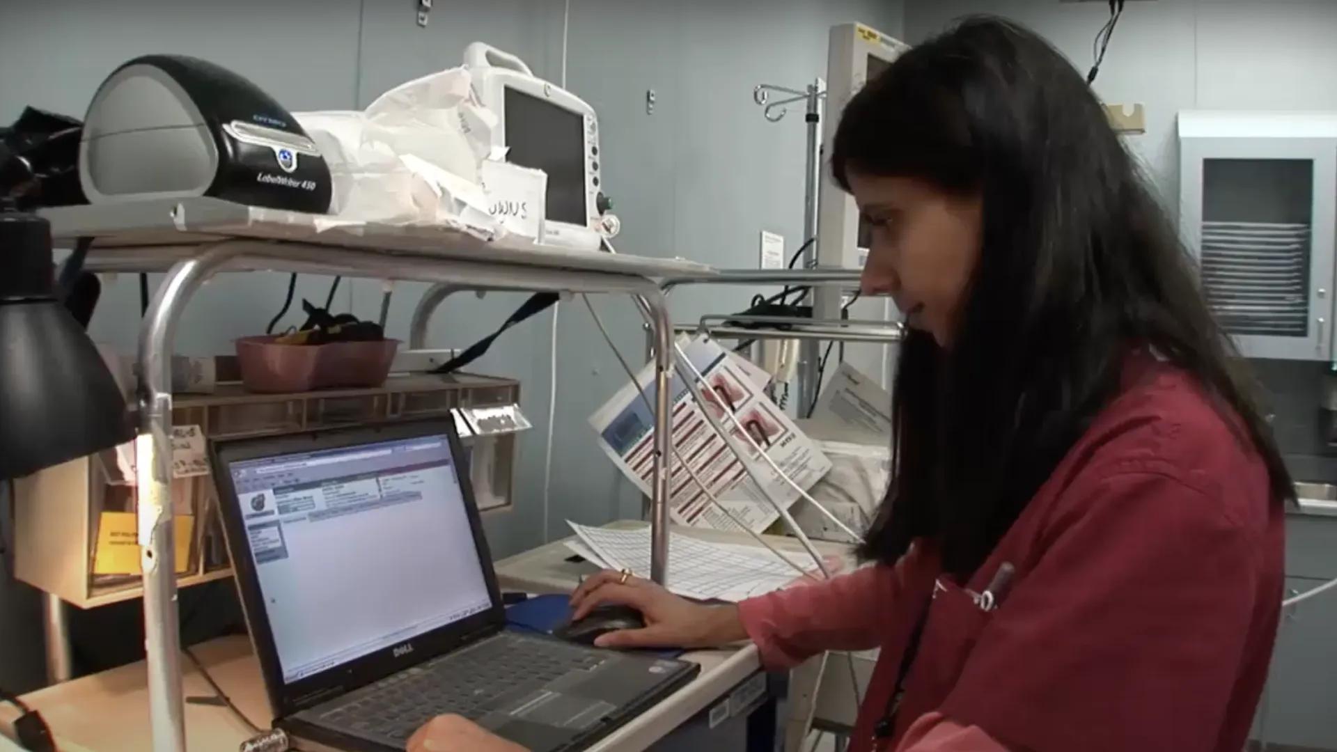 A woman in a maroon top is focused on a laptop in a clinical setting. 