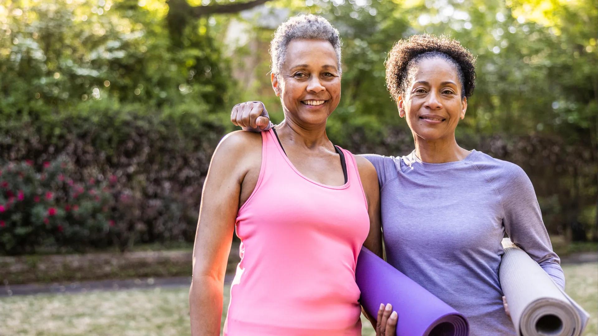 two Black women standing next to eachother, one in a pink tank top, one in long sleeve purple shirt, with yoga purple matts