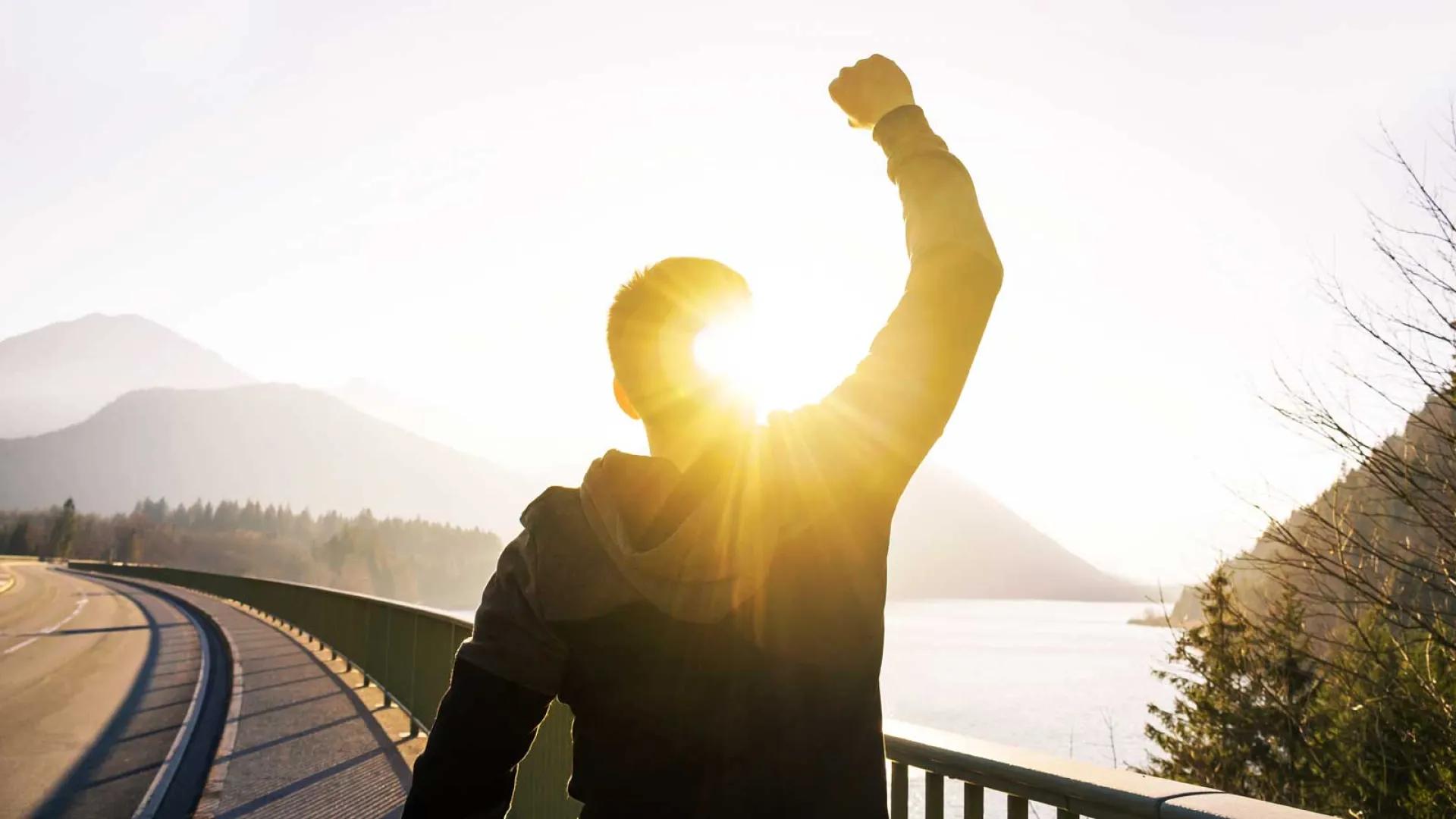 Man silouetted on a scenic track pumping his fist while facing the sunset, water and the mountains