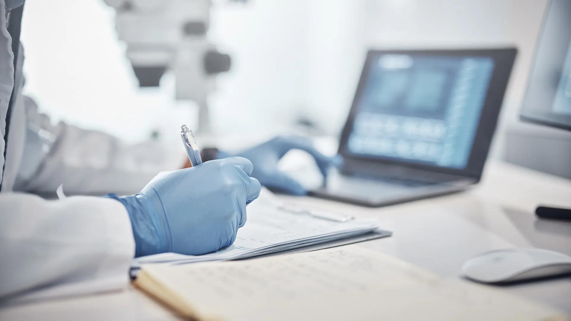 Person in lab coat and gloves writing notes and looking at a laptop on their desk