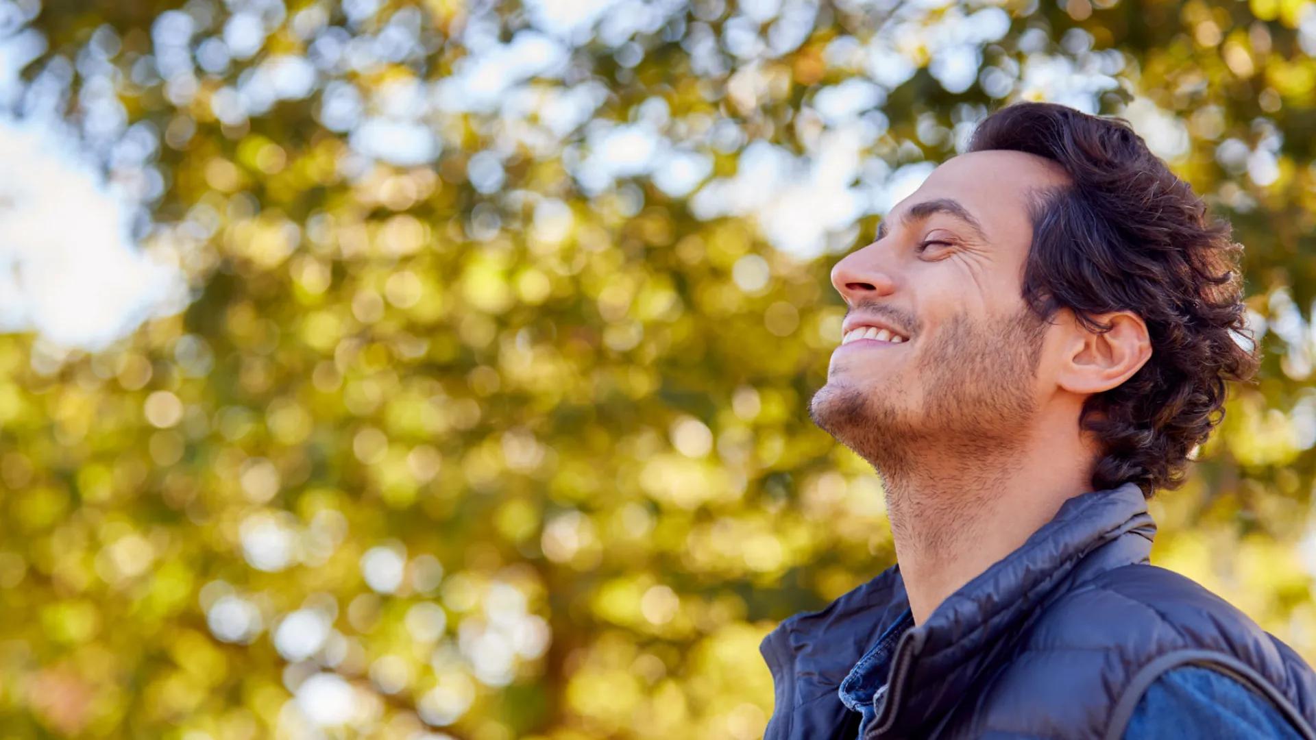 Man with eyes closed smiling. Trees full of leaves behind him.