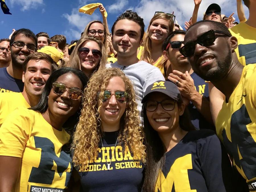 Group of smiling students in maize and blue at U-M football game on a sunny day
