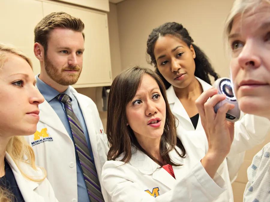 Group of doctors in white coat looking on as one doctor holds an instrument up to a patient's face
