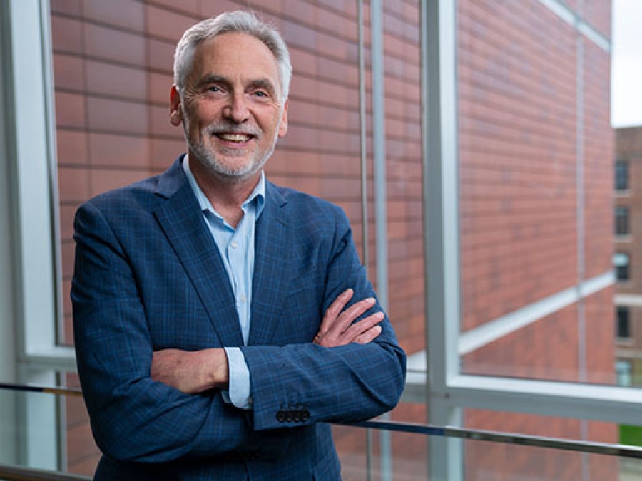 Smiling photo of Dr. Paulson standing in front of a large window. He is wearing a blue jacket and shirt, and a brick wall is visible behind him.