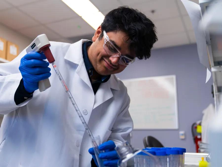 A researcher wearing a lab coat, safety glasses, and blue gloves smiles while using a pipette in a laboratory setting.