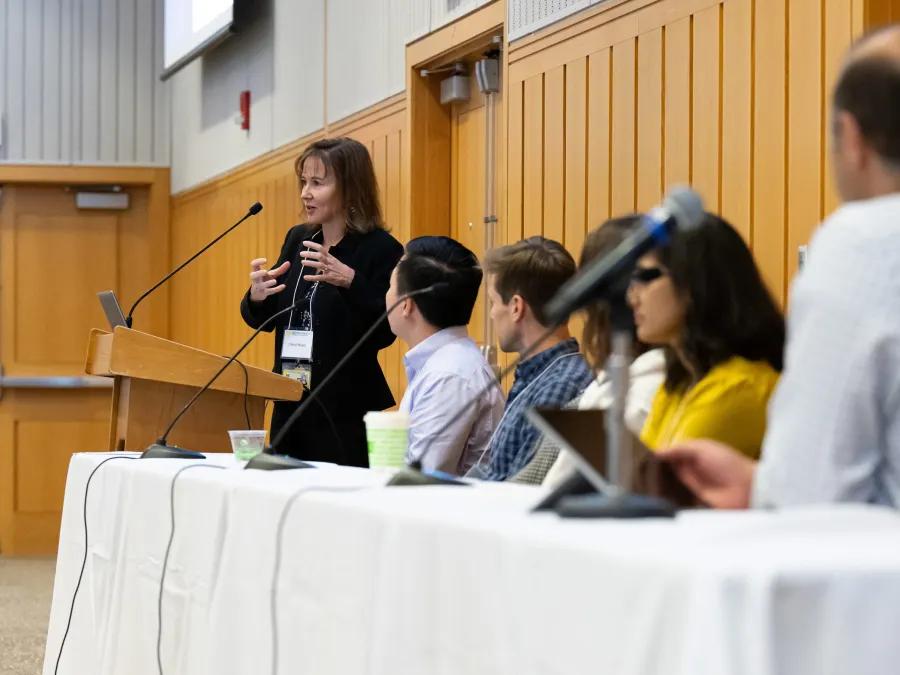 A panel discussion at a symposium featuring a speaker standing at a podium, gesturing while addressing the audience.