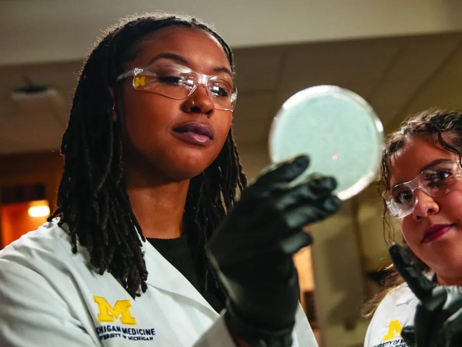 Two researchers in white coats and safety glasses. One is holding up a petri dish and the other is pointing at it.