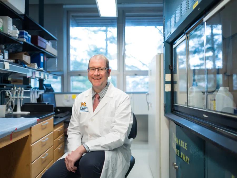 U-M's Dr Dan Wahl seated at lab bench in white coat, smiling 