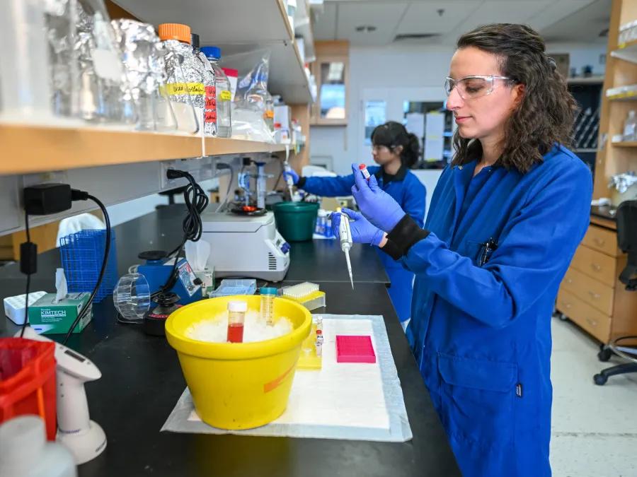 A woman in a blue lab coat wearing safety goggles and pipetting.