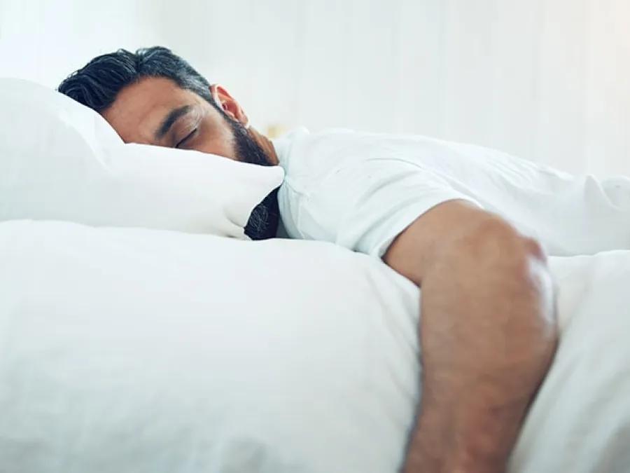 A man with a beard lies peacefully asleep on a bed, surrounded by soft white bedding.