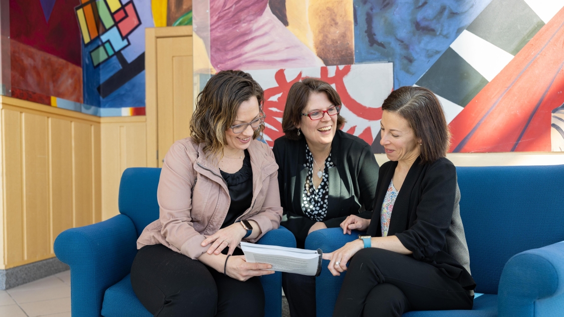  women sit on couch together and laugh while reading papers 