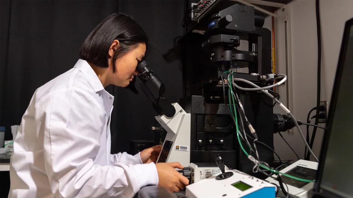 woman in a laboratory is leaned over while operating a microscope 