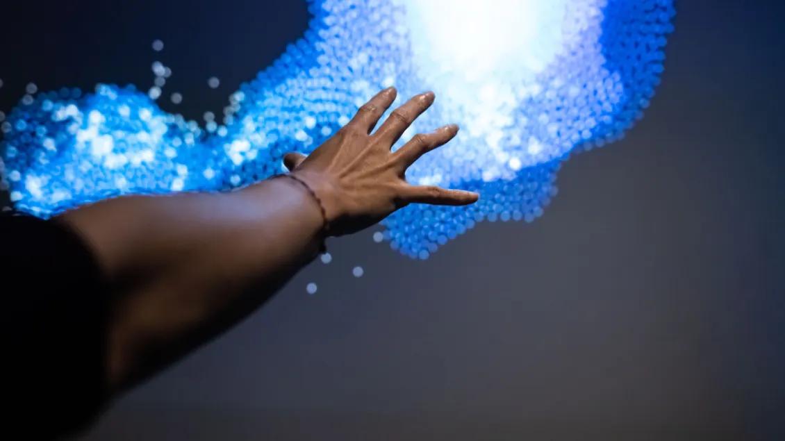A hand reaches towards a dynamic, abstract cluster of blue and white digital particles against a dark background, evoking a sense of interaction and technology.