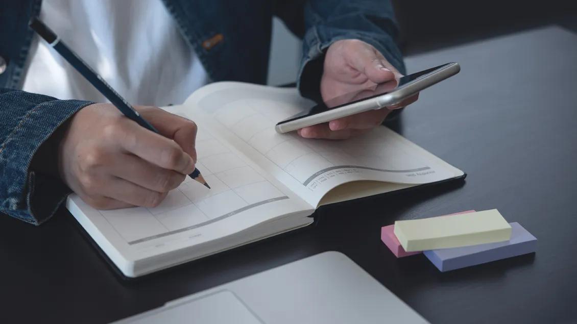 close-up of person writing in calendar notebook while looking at phone