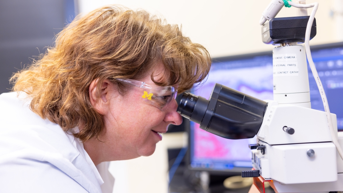 Woman in white lab coat and glasses looking through a microscope