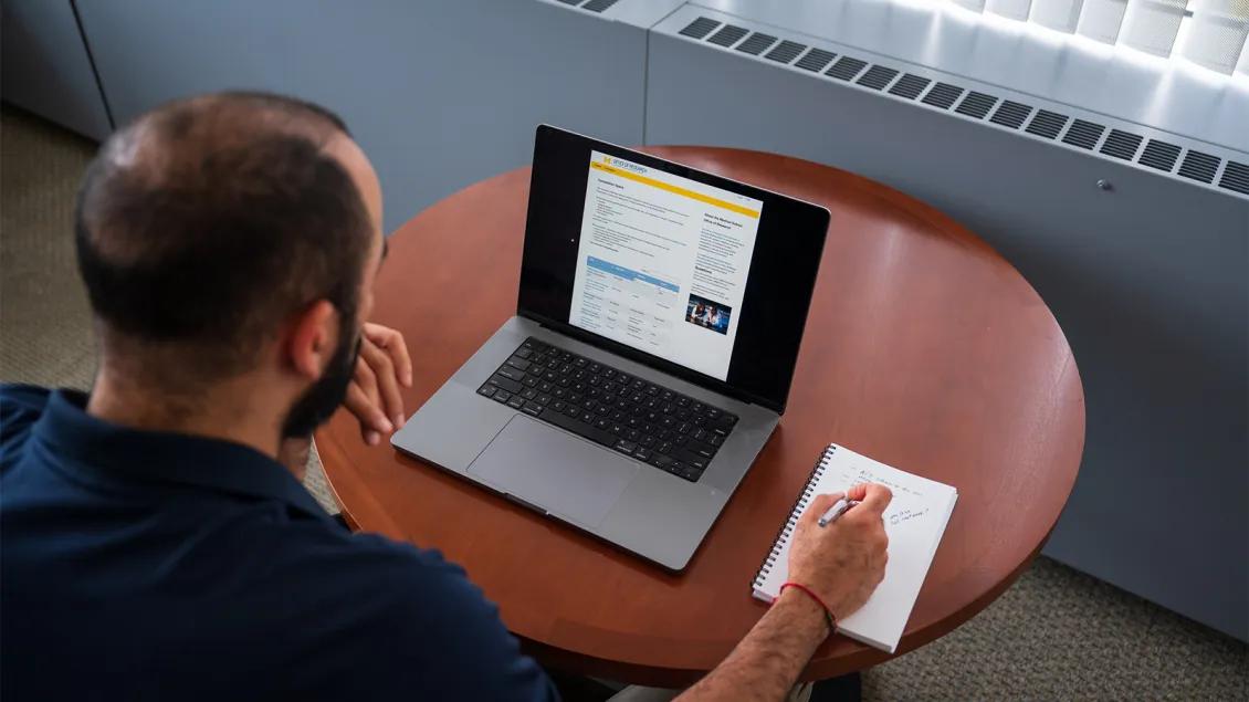 man looking at computer screen and writing in notebook