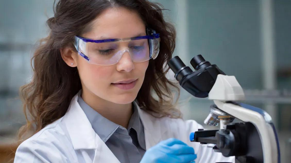 A scientist in a lab coat and safety goggles examines a sample under a microscope.