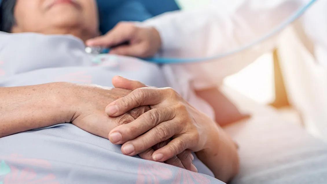 Elderly person lying in bed with hands clasped on chest, receiving a health check-up by a medical professional.