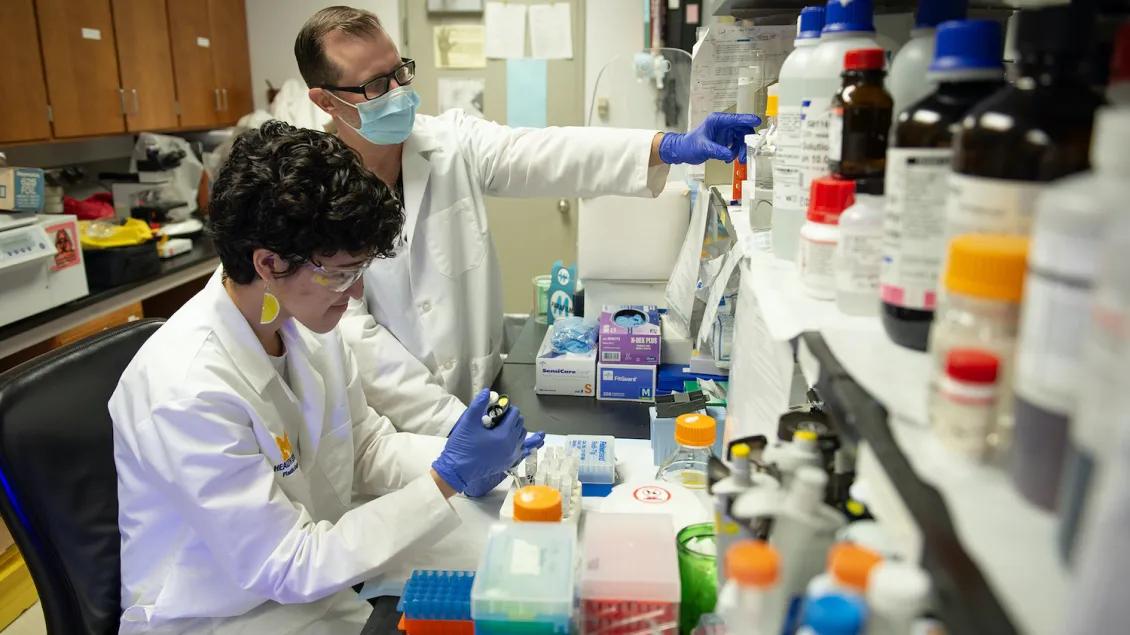 Two scientists in a lab, wearing white coats and gloves. One is seated, using equipment; the other stands, observing.