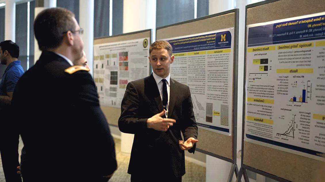 A man in a suit explains research on a poster board to a listener at a professional conference.