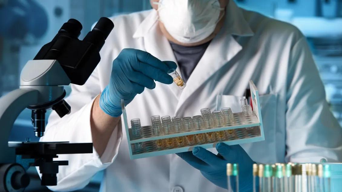 A masked research pulls a test tube from a tray