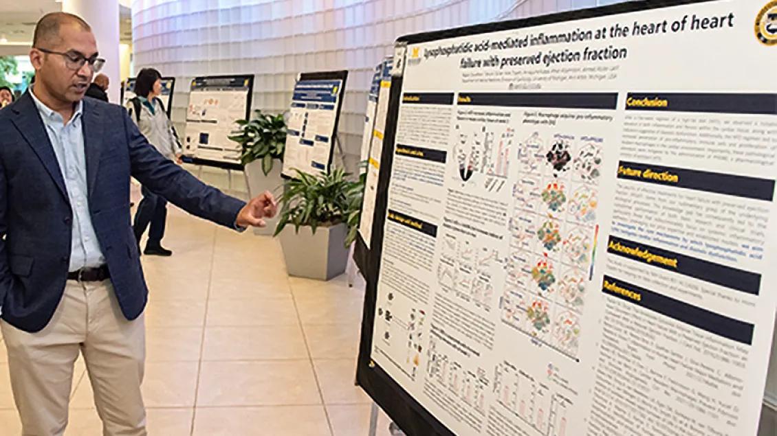 A man in a suit gestures toward a scientific poster at a conference.