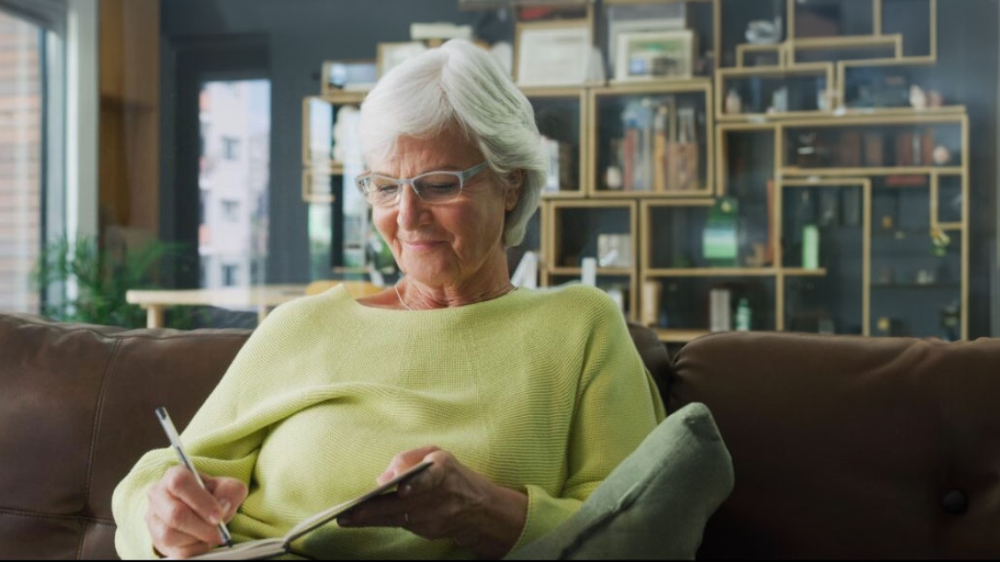 A white haried woman writes in her journal while sitting on a leather couch