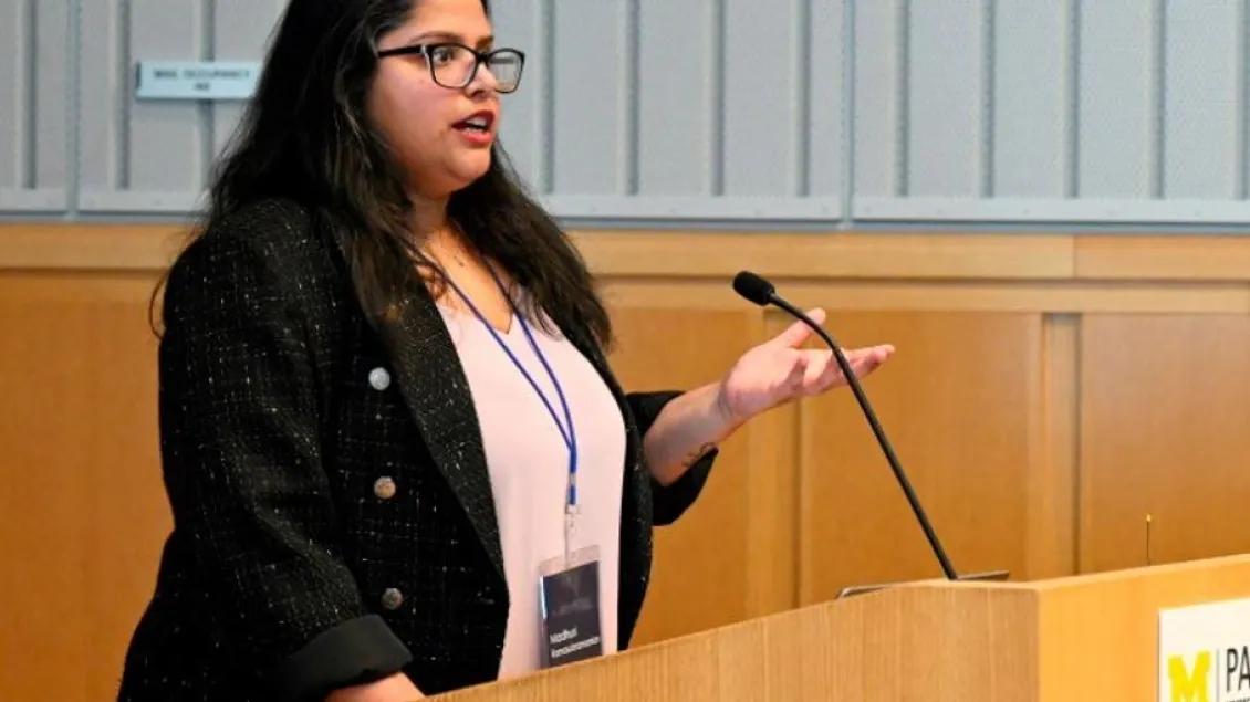 A woman speaks confidently at a wooden podium, wearing glasses and a dark blazer.