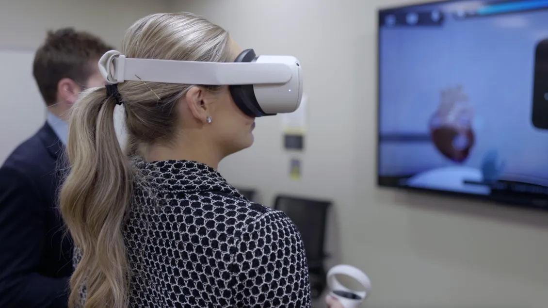 A person wearing a virtual reality headset stands in front of a screen displaying a virtual environment