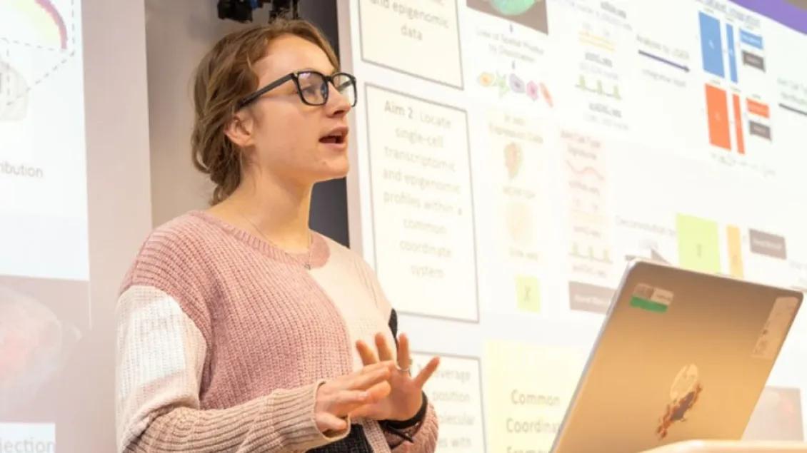 A woman wearing glasses and a multi-colored sweater gives a presentation in front of two large screens.
