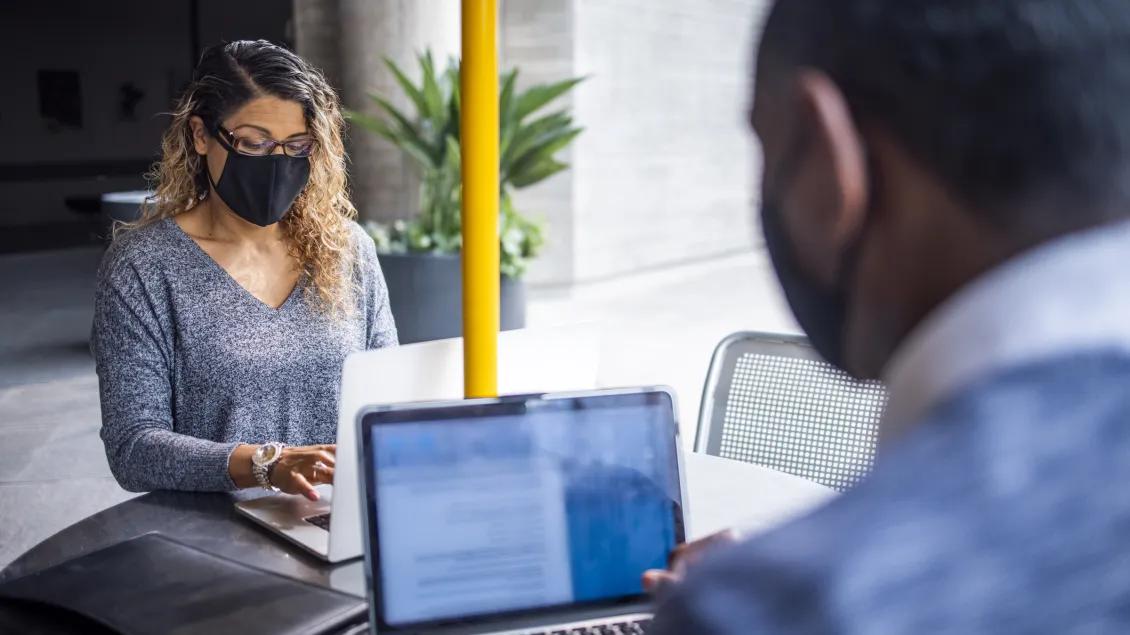 Two people wearing masks sit at an outdoor table with laptops.
