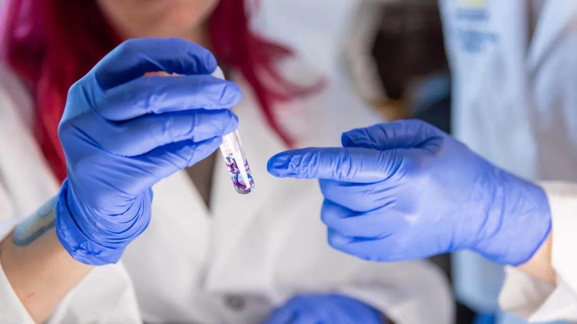 Two gloved hands holding and examining a test tube with a colorful liquid, with one person pointing at the tube for emphasis.