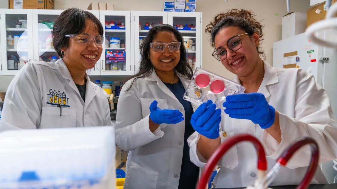 Three researchers in lab coats and safety glasses smiling as they examine a sample in a laboratory setting, with one holding a petri dish and the others observing.
